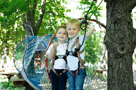 冒险公园里的两个可爱男孩正在攀岩或在绳路上越过障碍物，拥抱并竖起大拇指