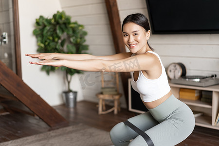 可爱的亚洲健身女孩在家锻炼，腿上绑着弹力绳蹲下，锻炼身体健康