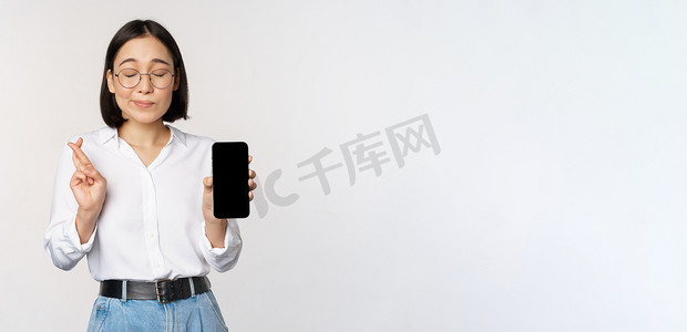 app展示界面摄影照片_充满希望的年轻亚洲女性，展示手机屏幕、应用程序界面和手指交叉，希望某物、许愿或付款，站在白色背景上