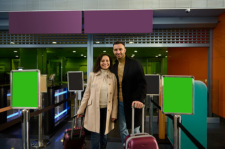 商务签到摄影照片_快乐的年轻夫妇，男人和女人，商业伙伴带着行李和手提箱站在登机柜台，在出发机场航站楼通过海关和护照检查。