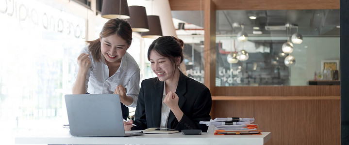 两位年轻的亚洲女商人在现代办公室里用笔记本电脑开心地微笑着，表现出工作成功的喜悦表情。