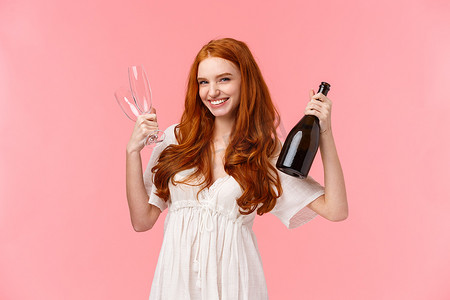 酒红色头发摄影照片_时髦漂亮的红头发女人和女朋友一起庆祝，一起玩乐，拿着一瓶香槟和两杯酒，邀请喝酒，面带笑容，开心，站着粉红色的背景