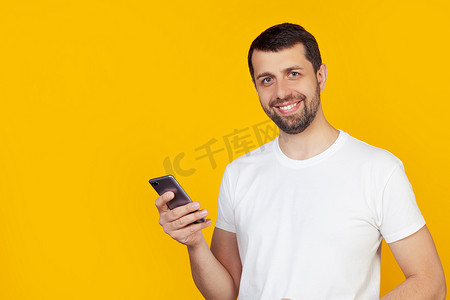 留着胡须、穿着白色 T 恤的年轻人正在使用智能手机，脸上带着快乐的脸站着，带着自信的笑容露出牙齿微笑。