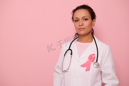 国际卫生组织摄影照片_自信的混血女医生肖像，身穿带粉红丝带的医用大衣，脖子上挂着听诊器，看着相机，在彩色背景中被隔离，复制空间。