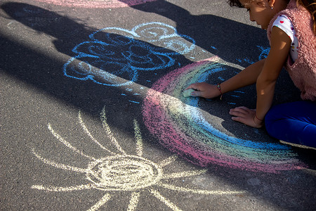 女孩在沥青上用粉笔画彩虹、太阳、汽车。