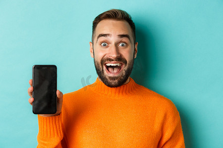 留着胡须的帅哥穿着橙色毛衣的特写，展示智能手机屏幕并微笑，展示在线促销，绿松石色背景