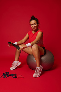 西班牙裔女运动员手持弹性健身带，在红色背景下锻炼后坐在健身球上。