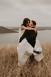 恩爱夫妻的婚礼，身穿礼服面纱、运动鞋和西装的新婚夫妇拥抱亲吻，在河上山田里的高高的草地上旋转