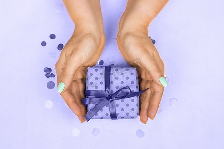 美甲贴纸新年摄影照片_2022 年左右，女人的手上涂着蓝色柔和的美甲，手里拿着紫色背景上带蝴蝶结的礼物。节日、礼物、惊喜、生日、新年和圣诞节的概念