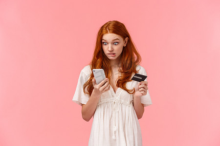 忧心忡忡、笨拙的可爱红发女孩犯了错误，在购物时不小心浪费了所有男朋友的钱，看着智能手机显示屏，拿着信用卡，看起来很内疚