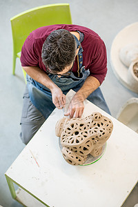 明亮陶瓷工作室中穿着围裙的陶艺家用生粘土雕刻雕像的俯视图。