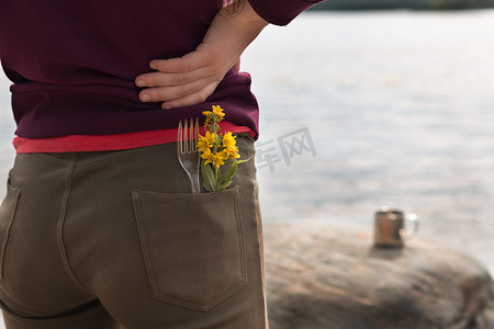 穿着棕色牛仔裤和紫色运动衫、口袋里插着叉子和黄色花朵的英俊女游客站在河岸上，欣赏夏季的山景。