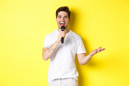 站立的麦克风摄影照片_快乐的男艺人表演，用麦克风说话，发表演讲或站立表演，站在黄色背景上