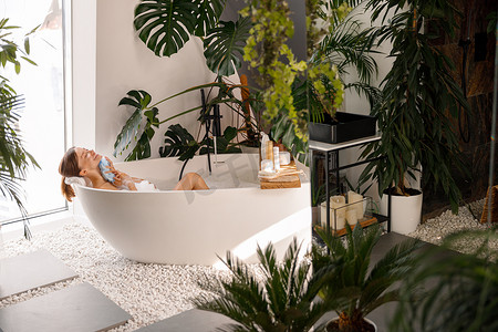 在装饰有热带植物的现代浴室内沐浴的放松的年轻女性