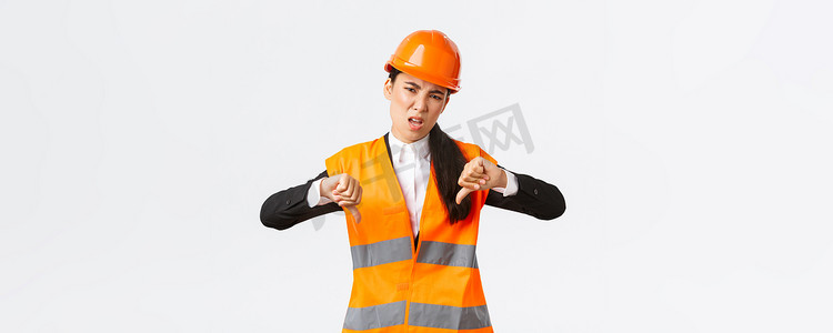 失望和不安的亚洲女总工程师检查糟糕的工作，表现出大拇指向下的失望，戴上安全头盔，抱怨建筑师的糟糕工作，白人背景