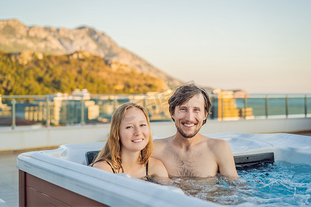 在绿色大山的背景下，享受快乐旅行时光假期生活的年轻无忧无虑的快乐微笑夫妇在热水浴缸中放松的肖像