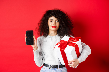 购物app摄影照片_一头卷发的漂亮女人，在空的智能手机屏幕上显示购物应用程序，拿着用节日盒子包裹的礼物，站在红色背景上