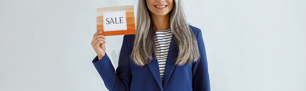 头发灰白的漂亮亚洲女士拿着浅色背景上写着“Sale”字样的卡片