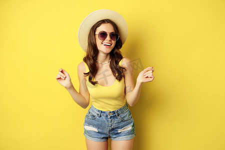 戴着草帽和背心的时尚女孩，准备迎接夏天，去度假，微笑着高兴，站在黄色背景上