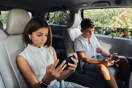 年轻的白人女孩和她十几岁的弟弟坐在小货车里专注于智能手机，孩子们在周末家庭自驾旅行中玩游戏