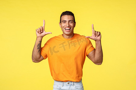 身穿橙色 T 恤、快乐开朗的年轻白人男子高兴地笑着，指着笑着宣传广告、提供建议、推荐下载应用程序或在商店购物、黄色背景