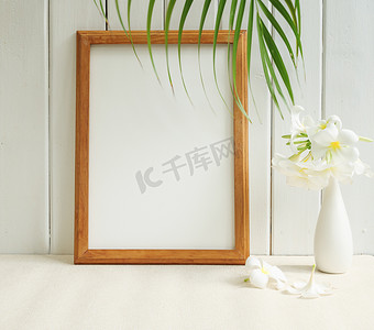 米色桌子上的现代白色花瓶中的模拟海报木框和美丽的鸡蛋花热带花卉，木墙背景上有绿色棕榈叶