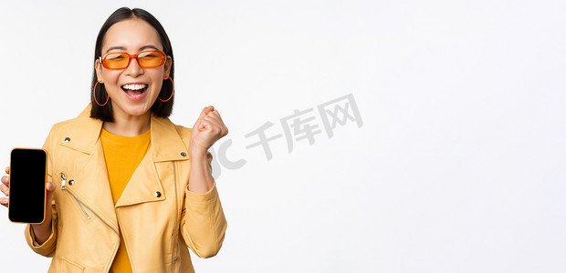 戴着太阳镜的快乐亚洲女孩，展示手机屏幕、智能手机界面，笑着，庆祝，站在白色背景上
