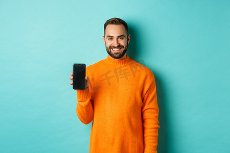 留着胡须的帅哥穿着橙色毛衣，展示智能手机屏幕，微笑着，在网上展示促销，绿松石背景