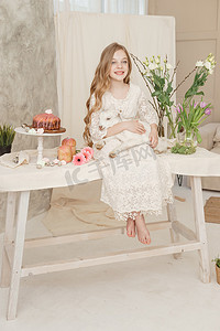 抱着花的女孩摄影照片_女孩坐在复活节餐桌旁，手里拿着蛋糕、春花和鹌鹑蛋，怀里抱着一个白人山羊。