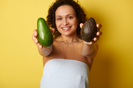 柔和的焦点放在裹着毛巾的模糊的半裸卷发微笑女人手中的成熟即食鳄梨水果上。