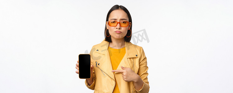 戴墨镜的悲伤亚洲女性肖像，手指指向手机应用程序界面，展示智能手机应用程序，站在白色背景上