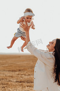 快乐的妈妈抱着她的孩子模仿飞行，年轻的妈妈在日落时分在城外和蹒跚学步的女儿玩耍