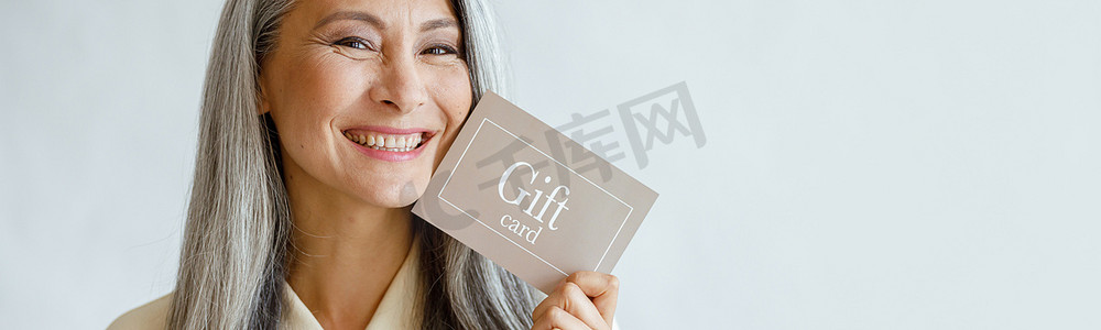 工作室里微笑着的成熟亚洲女士拿着浅灰色背景的礼品卡