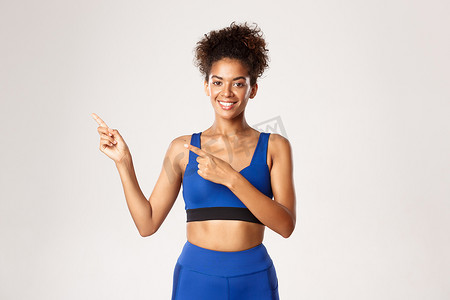 有魅力、健康的非洲裔美国健身女性，穿着蓝色运动服，手指指向左上角，微笑，展示标志，白色背景