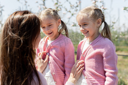 年轻的母亲在苹果园里为她的双胞胎女儿拉上粉色毛衣