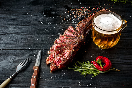 黑色木质背景中特写的烧烤干牛肉肋骨，配香料、蔬菜和一杯淡啤酒