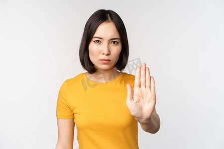 亚洲女性的肖像看起来严肃而愤怒，展示停止禁止手势、禁忌标志、禁止 smth，站在白色背景上的黄色 T 恤