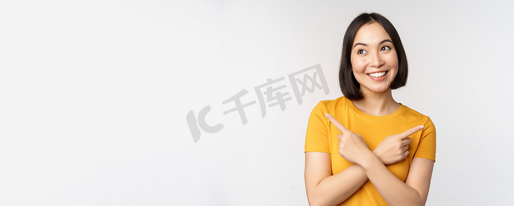 横板海报促销摄影照片_可爱的亚洲女孩手指横着，展示左右促销，两种选择，产品变体，站在白色背景上的黄色 T 恤