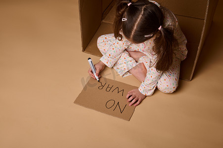 停止战争摄影照片_穿着睡衣的孩子在盒子里的纸板海报上写着“不战争”，躲避军事和政治冲突。