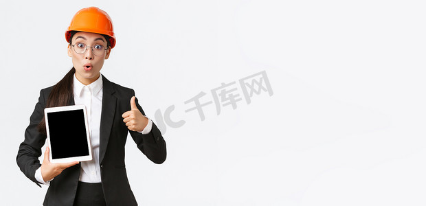 钱表情摄影照片_令人印象深刻和兴奋的亚洲女工程师、穿着西装和安全头盔的工业技术人员竖起大拇指，数字平板电脑屏幕上带着惊奇的表情，对利润图做出反应