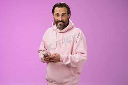 帅气的爸爸摄影照片_40 多岁时尚帅气、留着胡须的成年男子，穿着时髦的粉色连帽衫，拿着智能手机发短信，看起来很酷，很自信，心烦意乱，玩着很棒的新应用游戏，紫色背景
