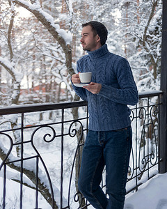 一个戴着帽子和蓝色毛衣的男人站在阳台上，端着一杯咖啡。
