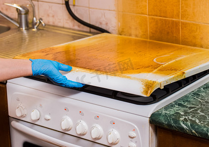 一位戴着家用橡胶手套的家庭主妇的手握着一个肮脏的煤气炉的盖子，上面有一层粘稠的涂层。