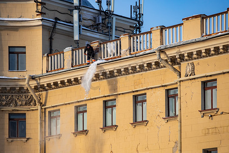 俄罗斯，圣彼得堡，2021 年 12 月 7 日：公用事业公司从屋顶除雪的工作，一个人扔掉屋顶的雪，他在屋顶结构上附有保险