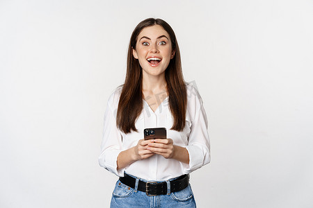 手机版k线摄影照片_带智能手机的年轻女性微笑着看着相机，使用手机应用程序、蜂窝技术和在线购物概念，白色背景