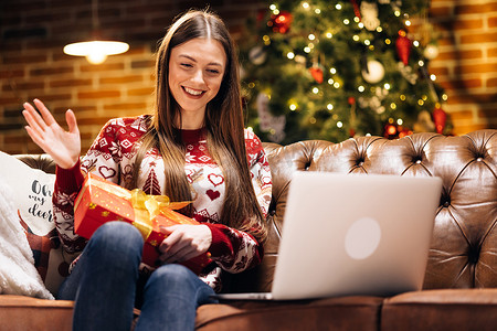 欢喜圣诞摄影照片_兴奋快乐的白人女孩戴着圣诞老人的帽子拿着圣诞礼盒，在电脑视频通话虚拟会议聊天中打开圣诞礼物与家人聊天，社交距离朋友坐在家里的沙发上