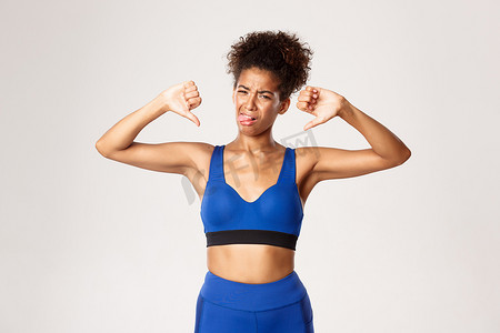 失望的非洲裔美国女运动员的摄影棚照片，身穿蓝色运动服，抱怨时竖起大拇指，因不喜欢而吐舌头，白色背景