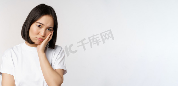 身穿休闲 T 恤的白色背景下，悲伤的亚洲女孩看起来心烦意乱、闷闷不乐、皱着眉头