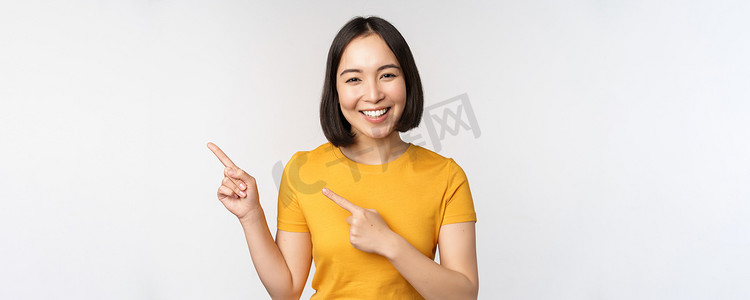 身穿黄色 T 恤、面带微笑的亚洲黑发女孩的肖像，手指向左，展示复制空间、促销交易、展示横幅，站在白色背景上