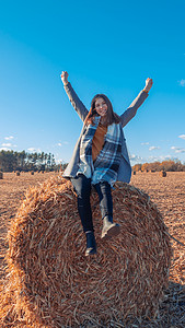 一个穿着灰色外套的欧洲女孩站在田野里，附近有一个更大的草捆，蓝天映衬着干草。